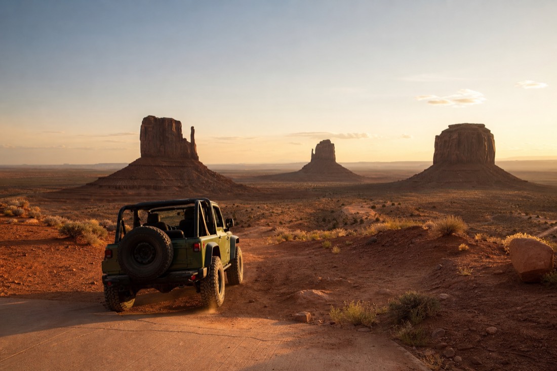 An open-air 4x4 jeep dropping off the paved apron onto red dirt below the park rim with the Mitten buttes ahead