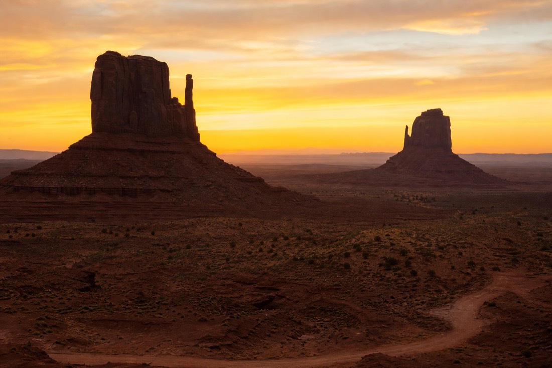 The two Mitten Buttes standing against a glowing sunrise sky, long shadows stretching across the sandy valley floor