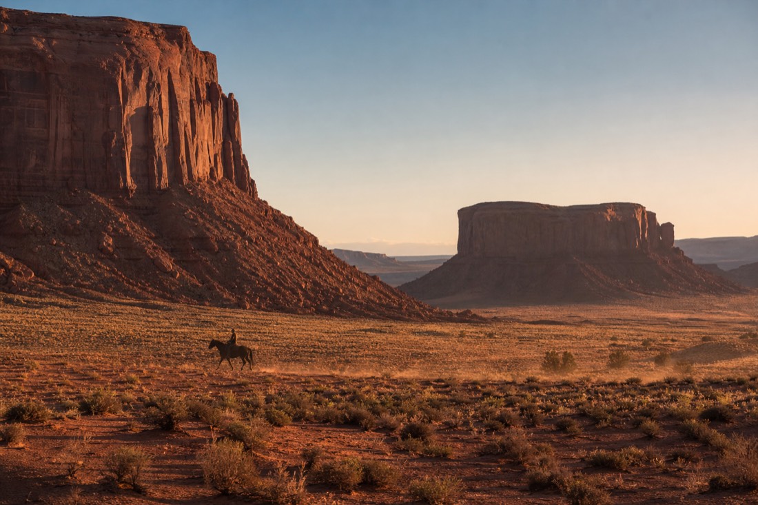 A lone silhouetted rider on horseback crossing the open valley floor between towering buttes at golden hour