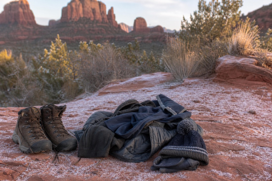 Warm winter clothing and boots laid out beside a frosted red dirt trail with buttes in the soft background