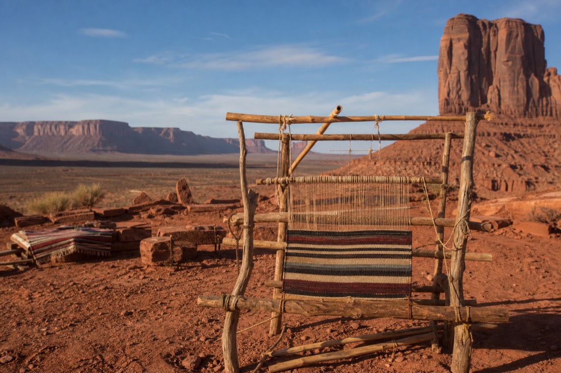 A traditional loom set up outdoors on the red desert floor with buttes rising in the background
