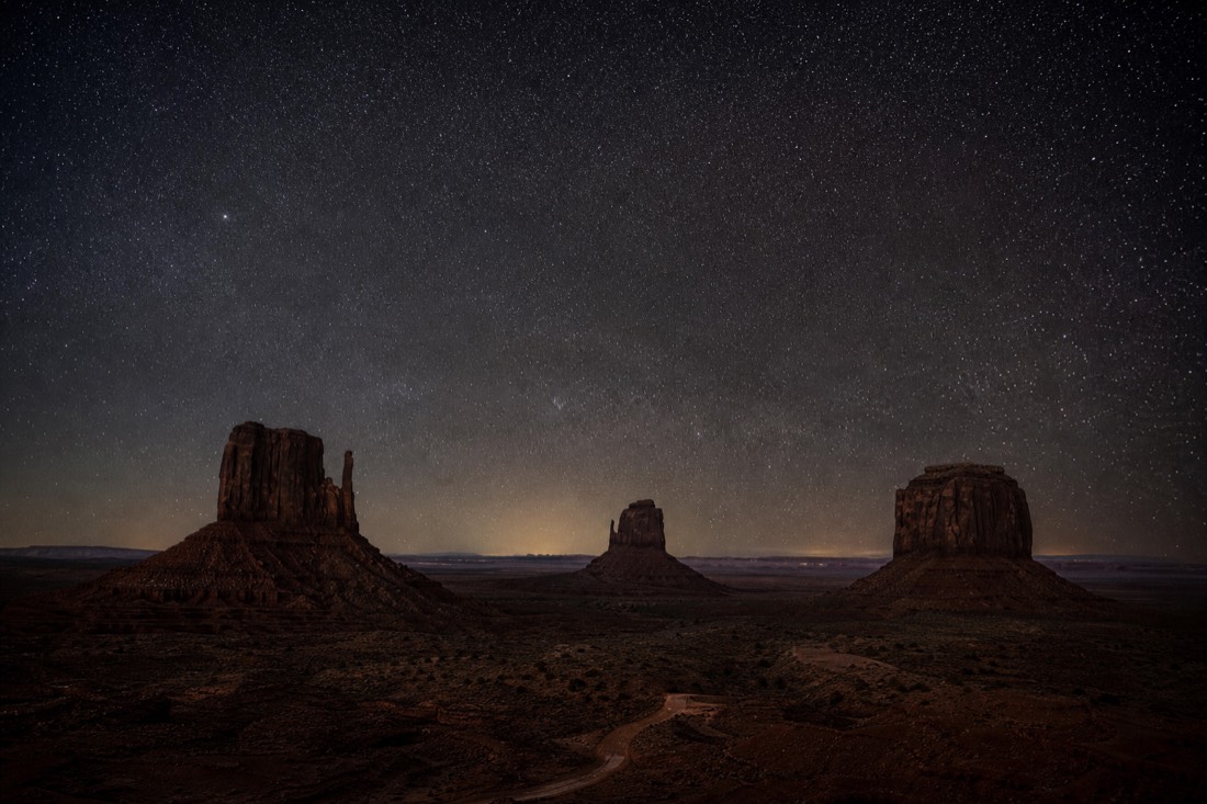 A vast dark sky filled with countless stars above the black silhouetted buttes of Monument Valley on a moonless night