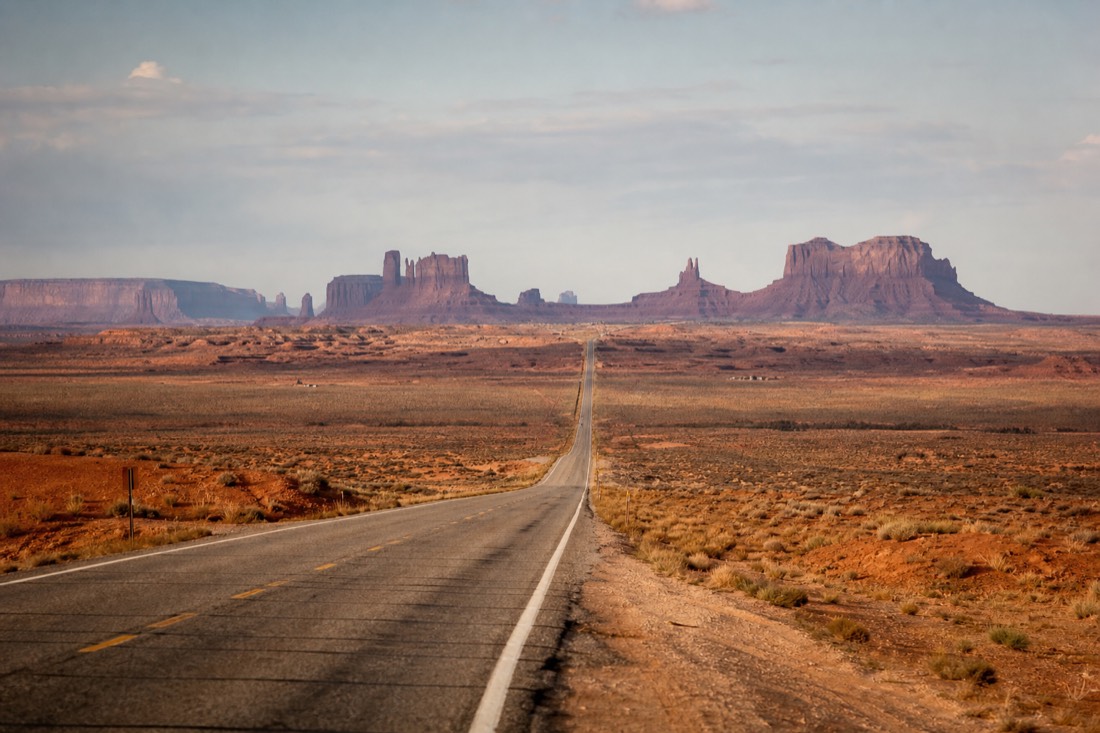 The long straight US-163 highway pointing directly at Monument Valley's buttes growing on the horizon, empty desert road ahead