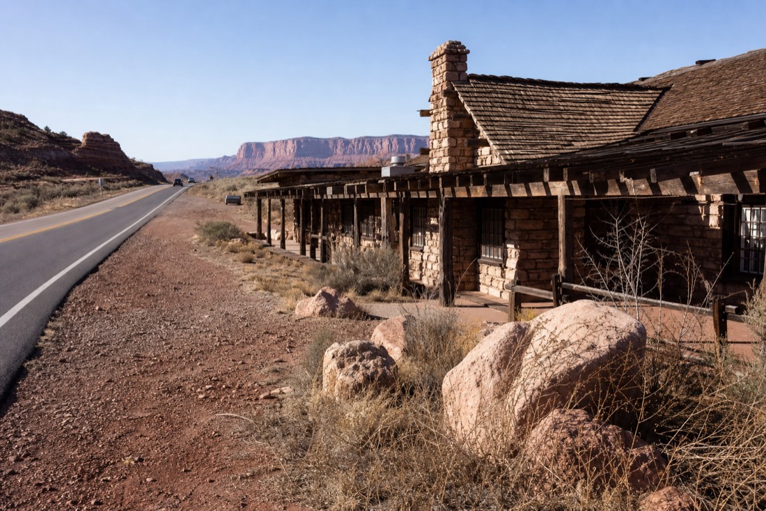 A historic stone-and-timber desert trading post beside a two-lane highway with red cliffs in the distance