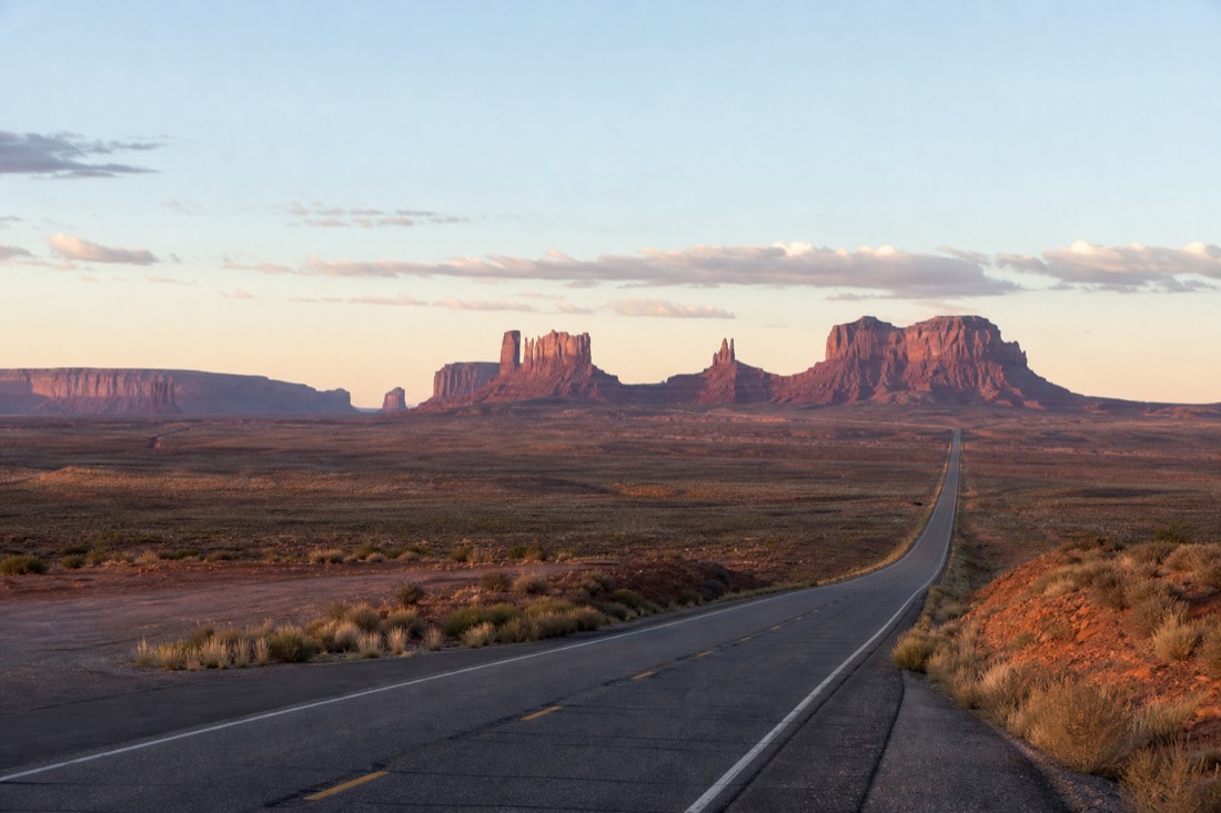 An open desert interstate stretching east toward distant red mesas at dawn, the long empty road to Monument Valley