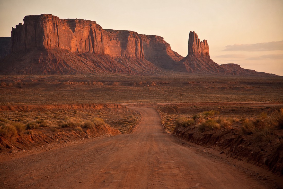 Vintage-style view down an empty dirt road toward towering buttes evoking a classic Western film setting at golden hour