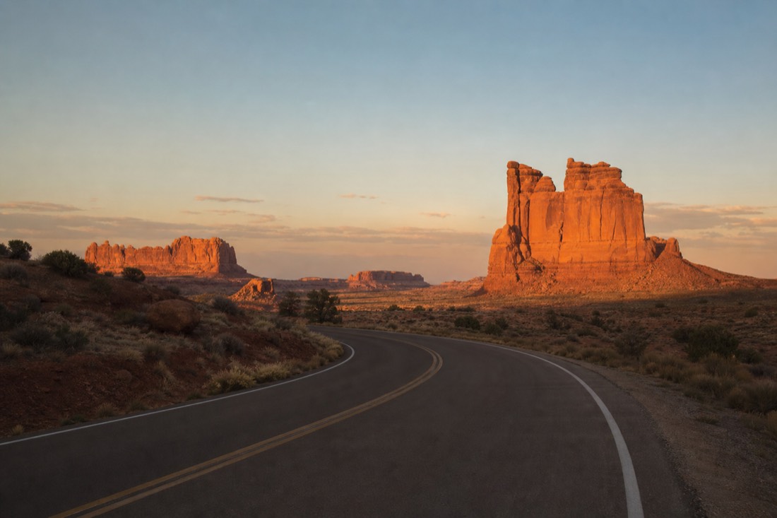 The park entrance road at golden hour with the buttes catching the last warm light before closing time