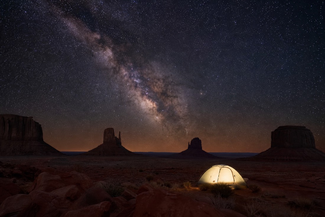 A small tent glowing under a brilliant Milky Way, Monument Valley buttes as dark silhouettes on the horizon